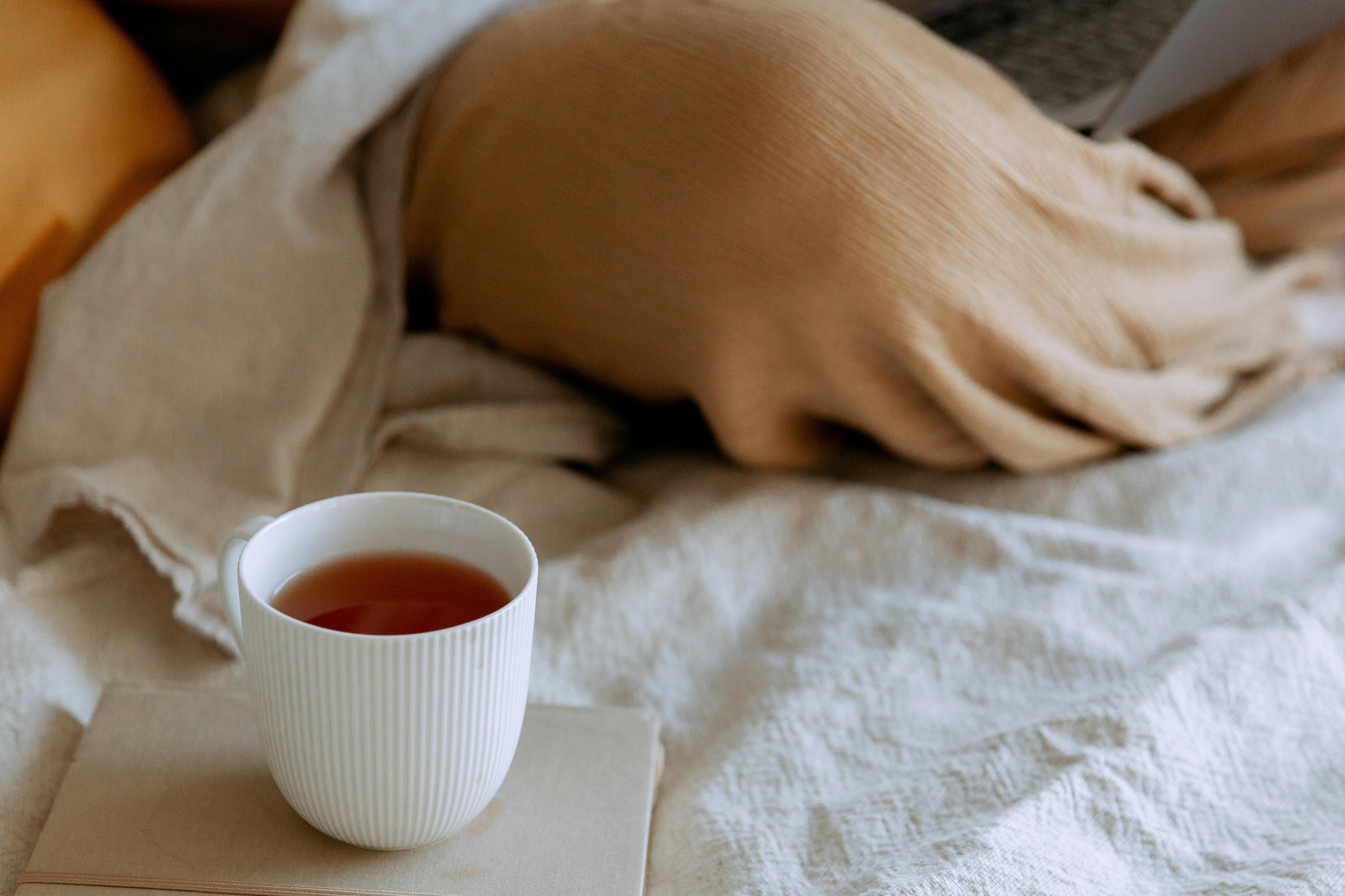 White mug with tea on a stack of books on a white blanket, with a person partially visible in the background.